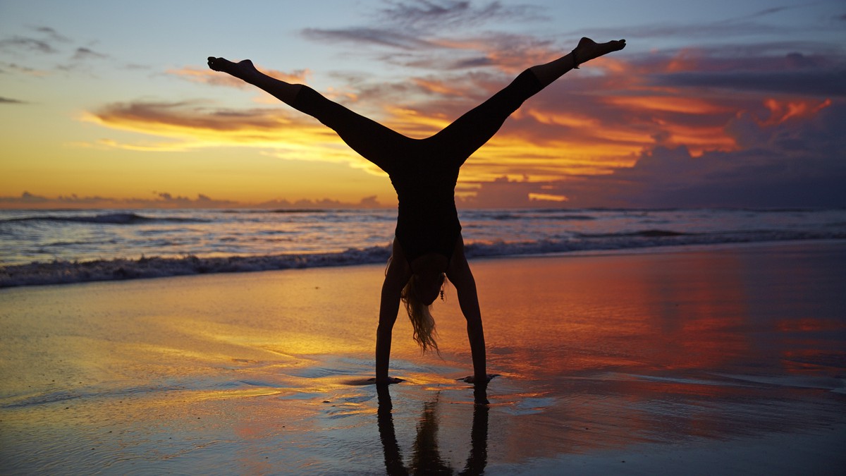 A silhouetted woman does a handstand with legs spread out in front of a beautiful sunset on a beach.