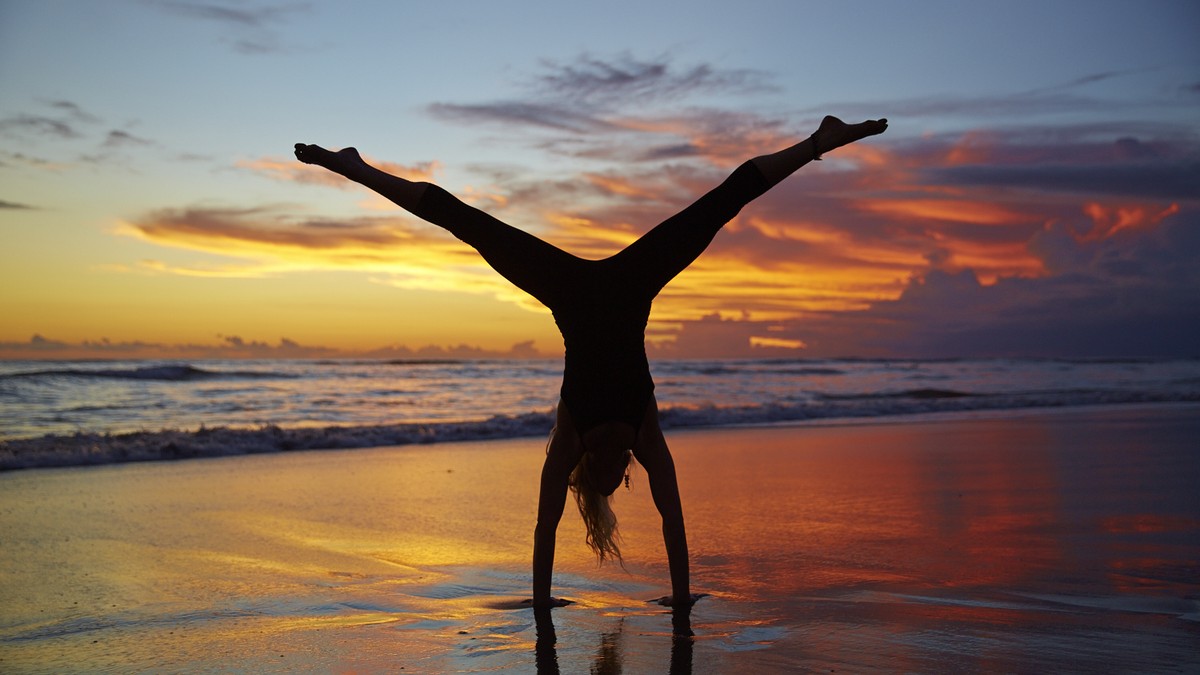 A silhouetted woman does a handstand with legs spread out in front of a beautiful sunset on a beach.