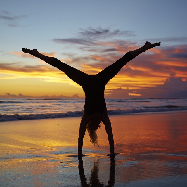 A silhouetted woman does a handstand with legs spread out in front of a beautiful sunset on a beach.