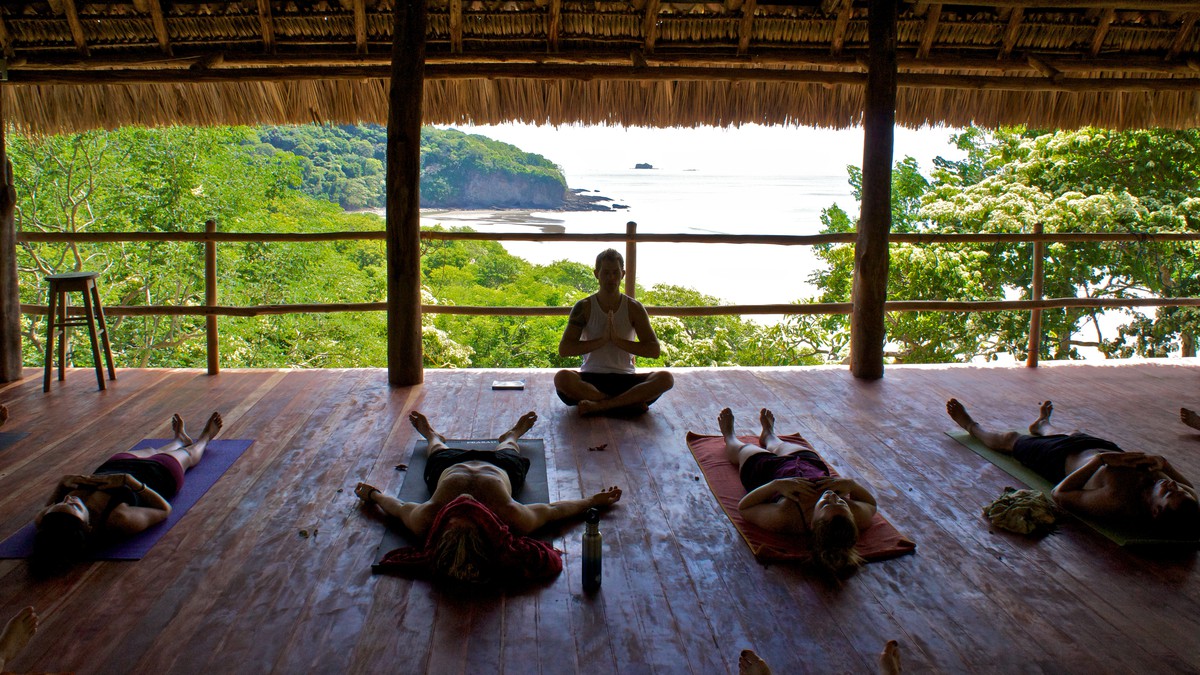A yoga instructor sits lotus in front of his students laying down on their backs. The yoga platform overlooks a huge beach, some jungle and the ocean.