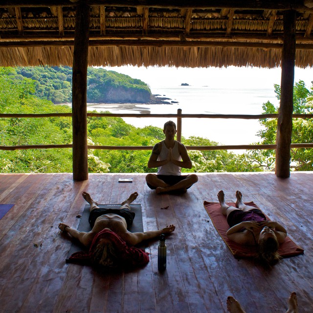 A yoga instructor sits lotus in front of his students laying down on their backs. The yoga platform overlooks a huge beach, some jungle and the ocean.